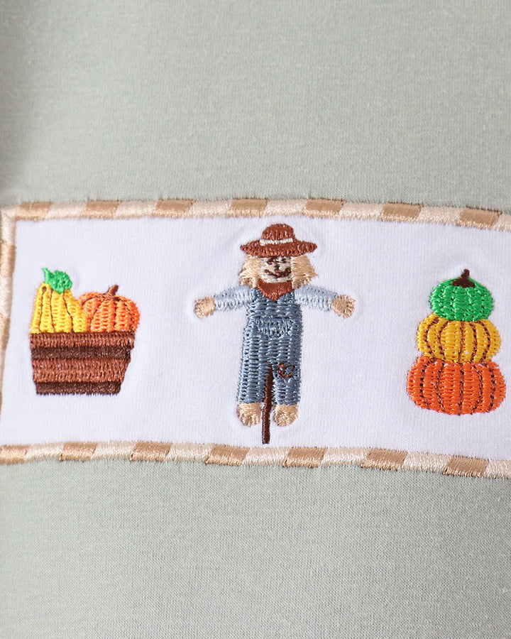 Close-up of a sage green garment featuring a white rectangular embroidered patch with a scarecrow in the center and two stacked pumpkins on either side.