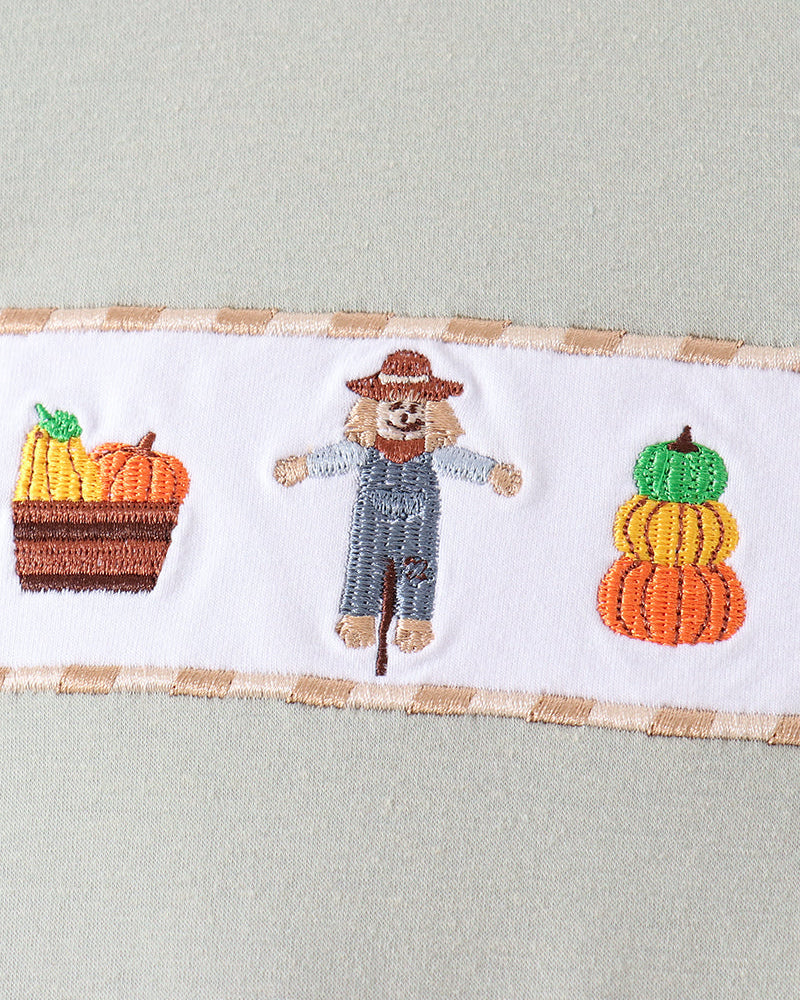 Close-up of a light sage green garment featuring a horizontal white band with autumn embroidery, including a scarecrow in blue overalls and baskets of pumpkins and corn.