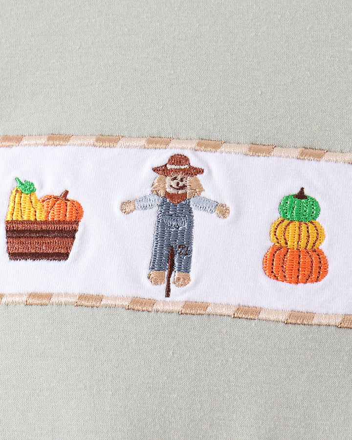 Close-up of a light sage green garment featuring a horizontal white band with autumn embroidery, including a scarecrow in blue overalls and baskets of pumpkins and corn.