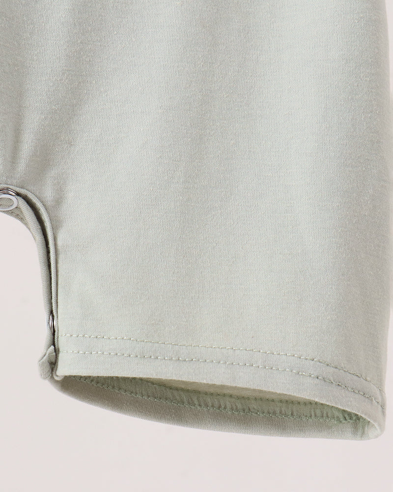 Close-up detail of a light sage green garment, showing a curved hem with a visible snap button closure and clean stitching on a plain light background.