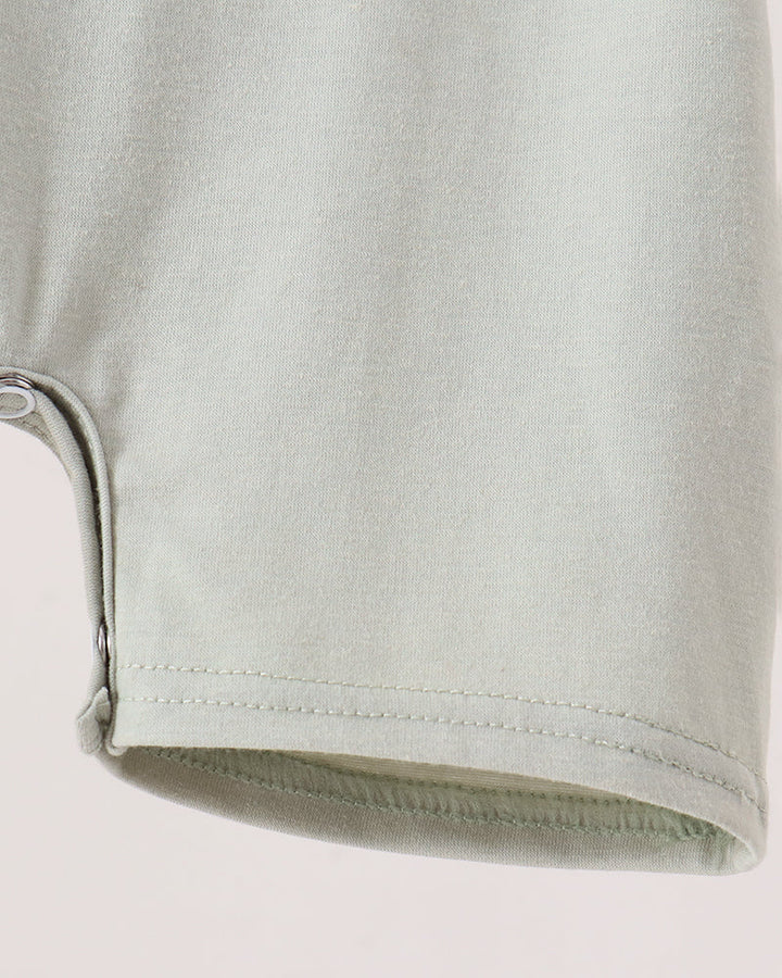 Close-up detail of a light sage green garment, showing a curved hem with a visible snap button closure and clean stitching on a plain light background.