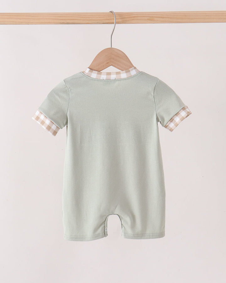 A baby short-sleeve romper in pale sage green, hanging on a wooden hanger from a light wood rod. The romper features a beige and white gingham patterned collar and sleeve cuffs, shown against a plain off-white background.
