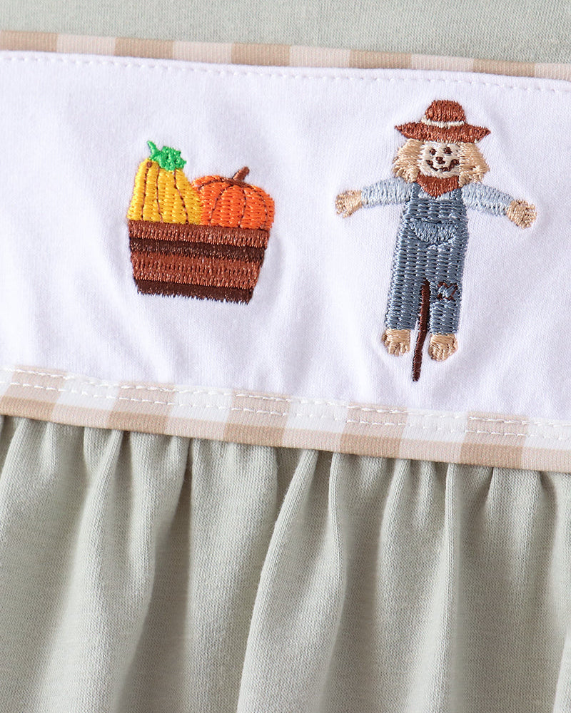 Close-up of a baby dress featuring a white horizontal band with embroidered autumn motifs: a basket of yellow and orange pumpkins and a scarecrow in blue overalls and a brown hat. The garment body is a soft sage green with a ruffled hem.