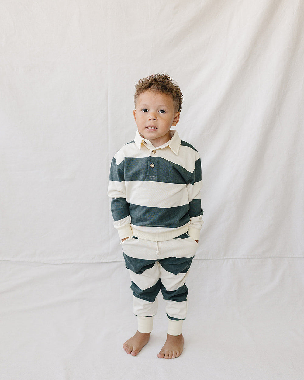 A toddler stands barefoot against a white fabric backdrop, wearing a long-sleeve collared rugby-style shirt and matching striped pants in dark green and cream.