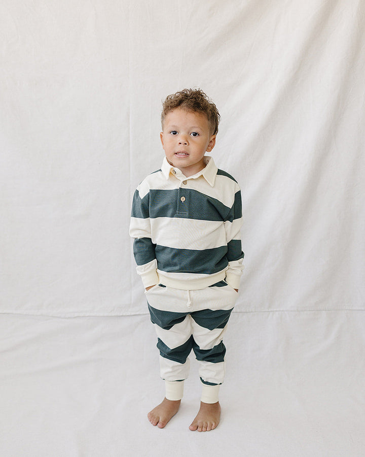 A toddler stands barefoot against a white fabric backdrop, wearing a long-sleeve collared rugby-style shirt and matching striped pants in dark green and cream.