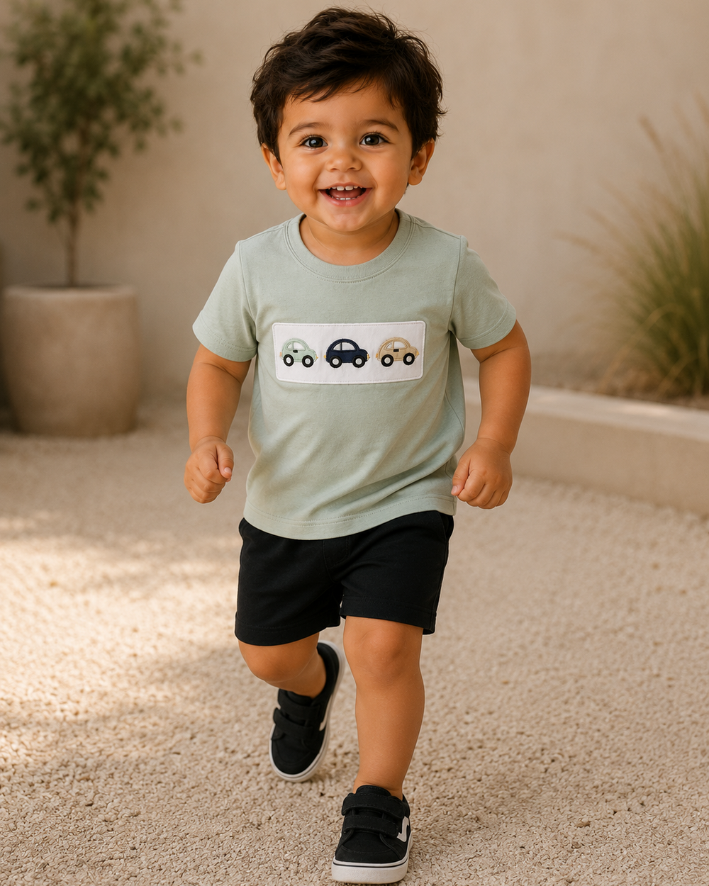 A toddler boy smiling and running while wearing a sage green short-sleeve t-shirt with a car graphic on the chest, black shorts, and black sneakers, set against a warm beige background with plants.