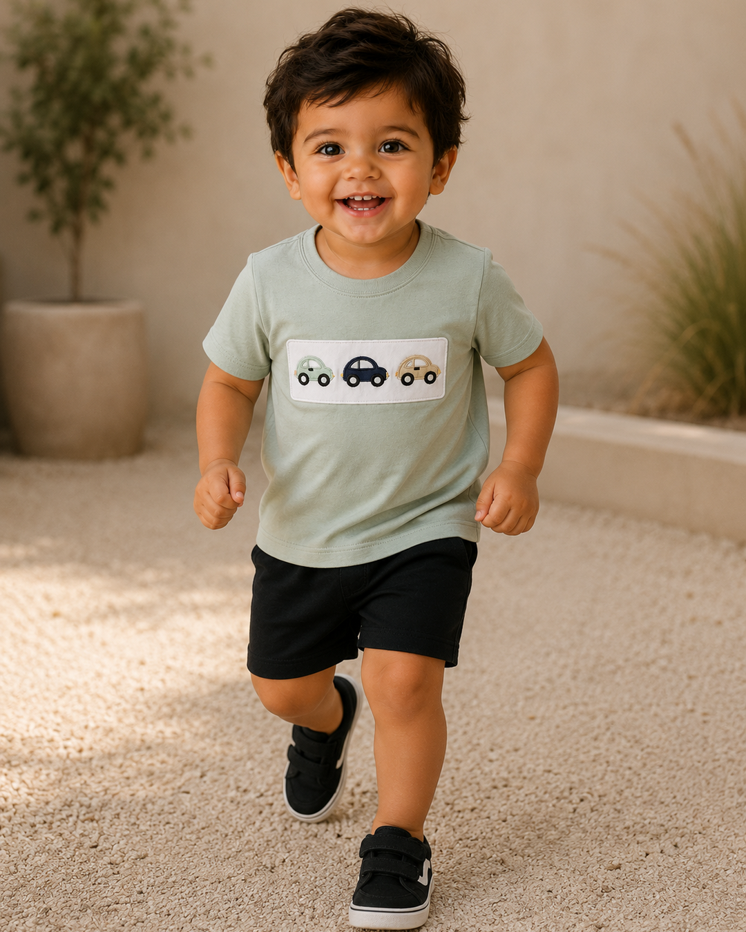 A toddler boy smiling and running while wearing a sage green short-sleeve t-shirt with a car graphic on the chest, black shorts, and black sneakers, set against a warm beige background with plants.