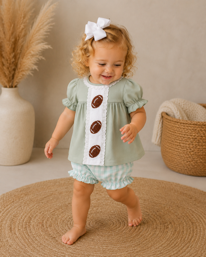 A toddler girl wearing a sage green and white football-themed romper with three brown football buttons, walking barefoot on a round jute rug in a styled nursery setting.