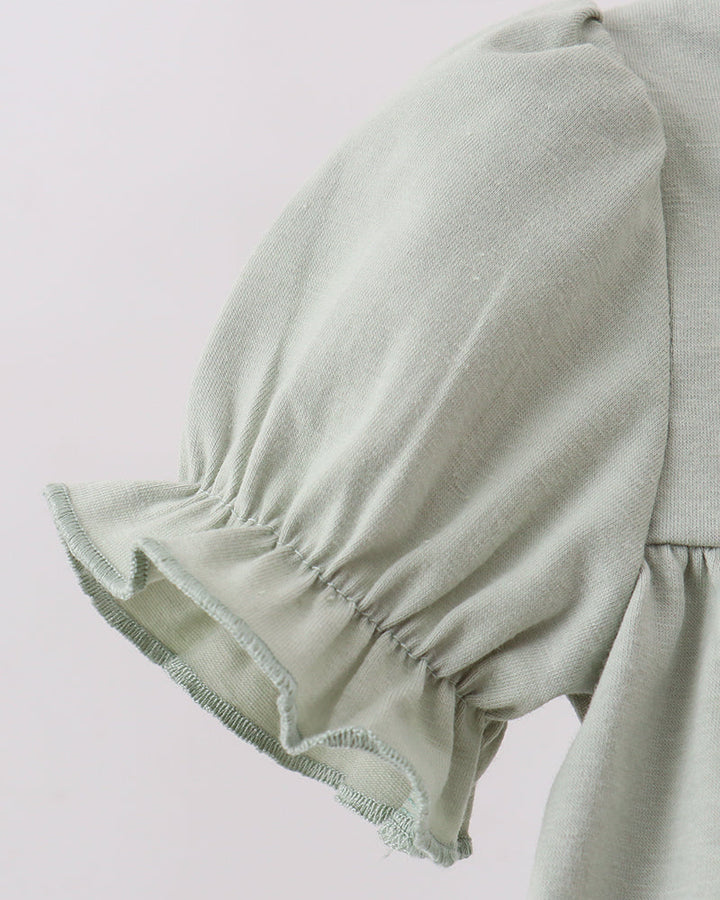 Close-up of a light sage green garment featuring a gathered puff sleeve with a ruffled hem and a matching solid fabric body.