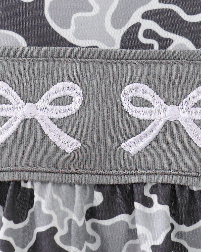Close-up of a gray ruffled trim with white embroidered bow motifs, layered over a gray, black, and white camouflage patterned fabric.