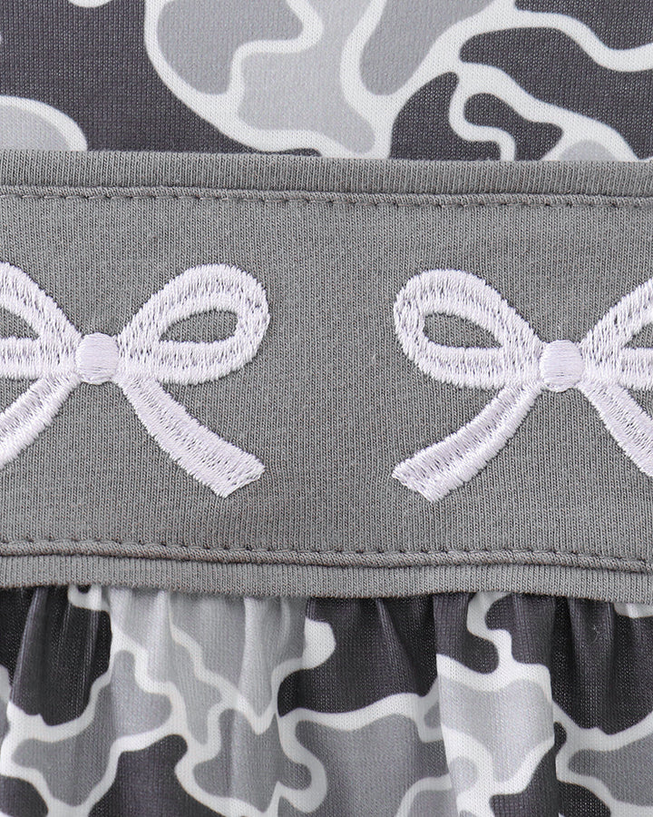 Close-up of a gray ruffled trim with white embroidered bow motifs, layered over a gray, black, and white camouflage patterned fabric.