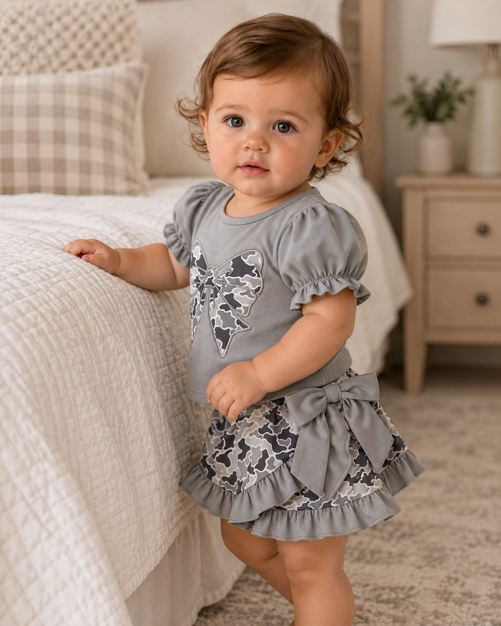 Baby girl in camo outfit is standing next to a bed.