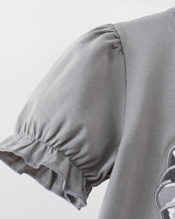 Close-up of a light gray short-sleeve top with gathered puff sleeves and a ruffled hem, showing part of a tonal graphic on the lower right.