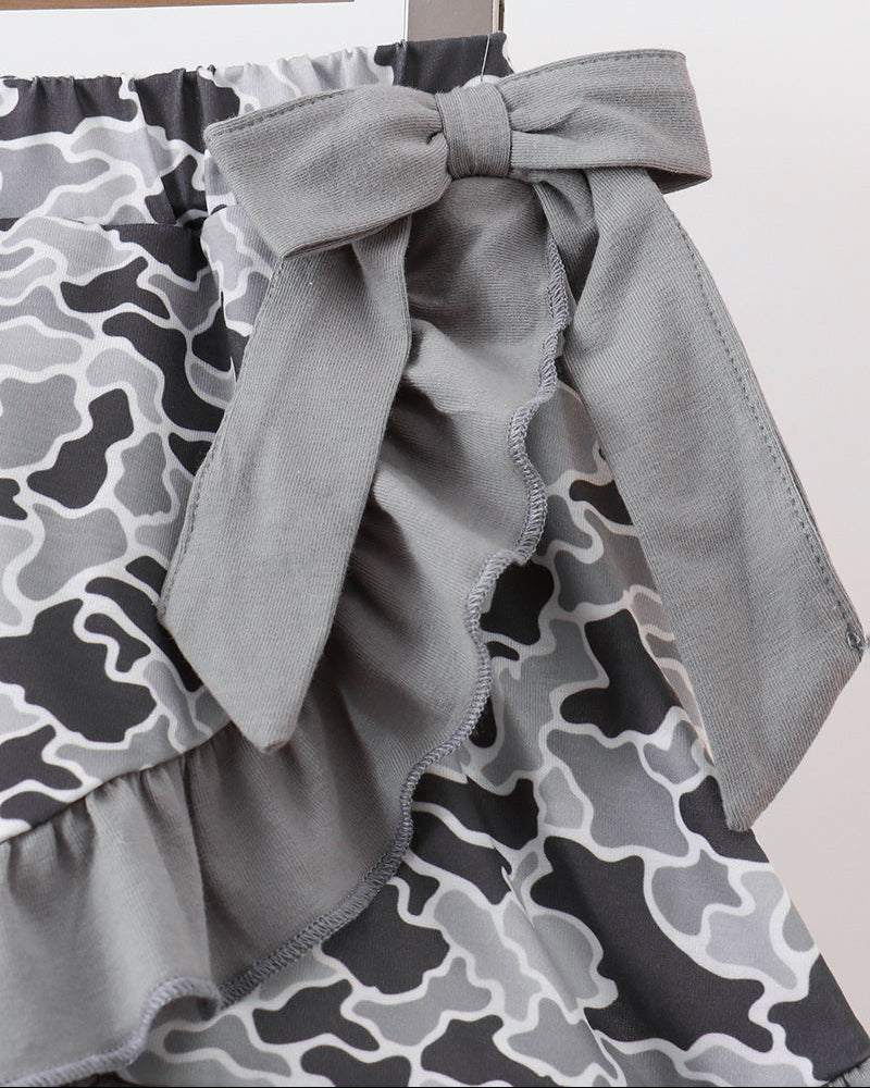 Close-up of a skirt with a gray ruffled bow and a black, gray, and white camouflage print, hanging on a wooden hanger against a plain light background.