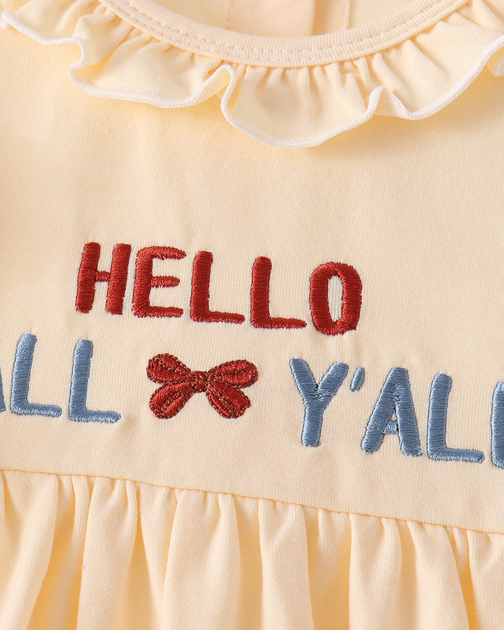 Close-up of a pale yellow baby dress with a ruffled neckline and embroidered text reading &