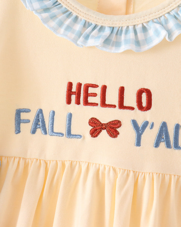 Close-up of a baby dress in pale yellow with a blue and white gingham ruffled collar and embroidered text reading &