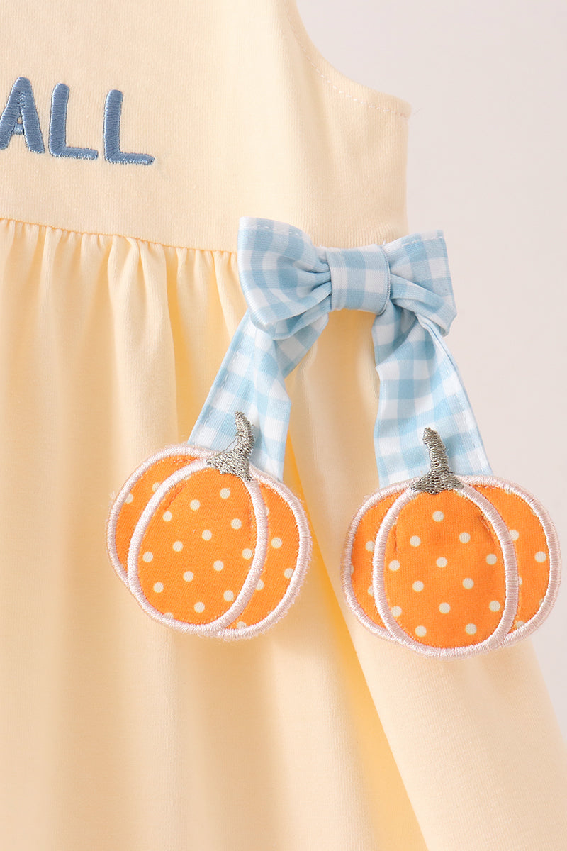 A close-up of a pale yellow dress with a blue gingham bow and two orange pumpkin appliques with white polka dots hanging from the front.