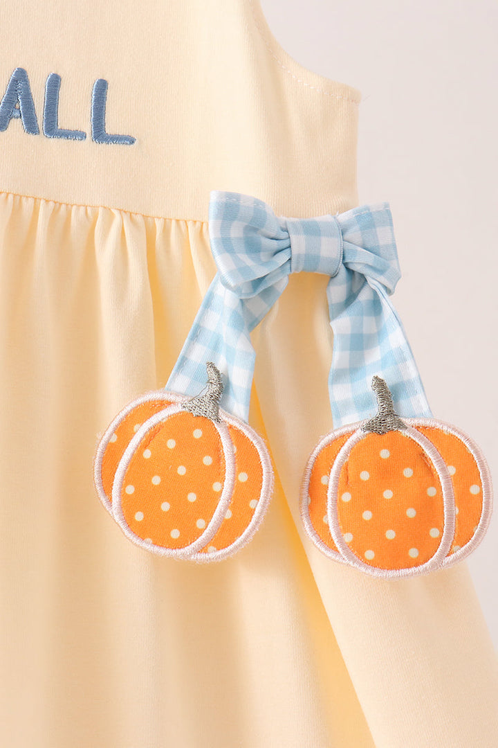A close-up of a pale yellow dress with a blue gingham bow and two orange pumpkin appliques with white polka dots hanging from the front.