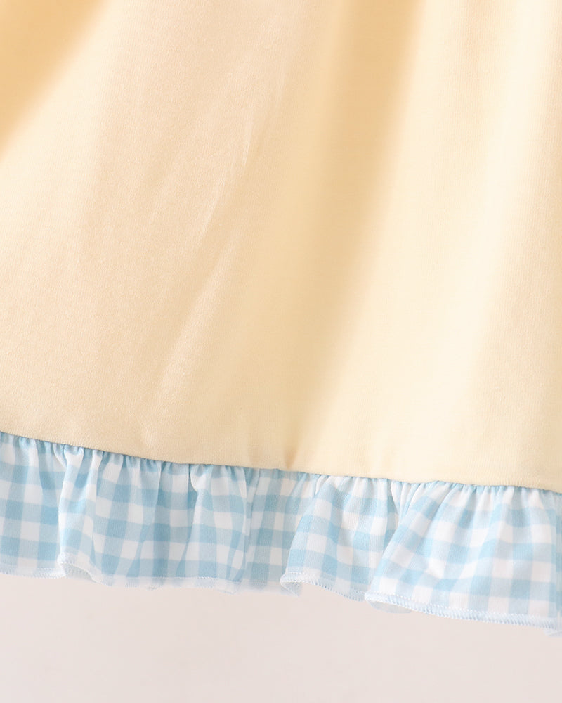 Close-up of a pale yellow fabric item with a ruffled hem made of light blue and white gingham check fabric, set against a plain light background.