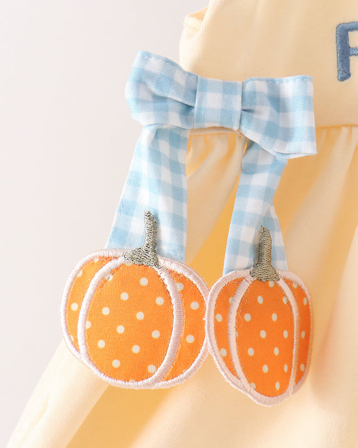 A close-up of a pale yellow garment with a light blue gingham bow and two orange pumpkin appliqués with white polka dots hanging from it.