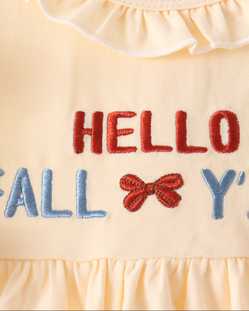 Close-up of a pale yellow baby dress with a ruffled neckline and embroidered text reading &