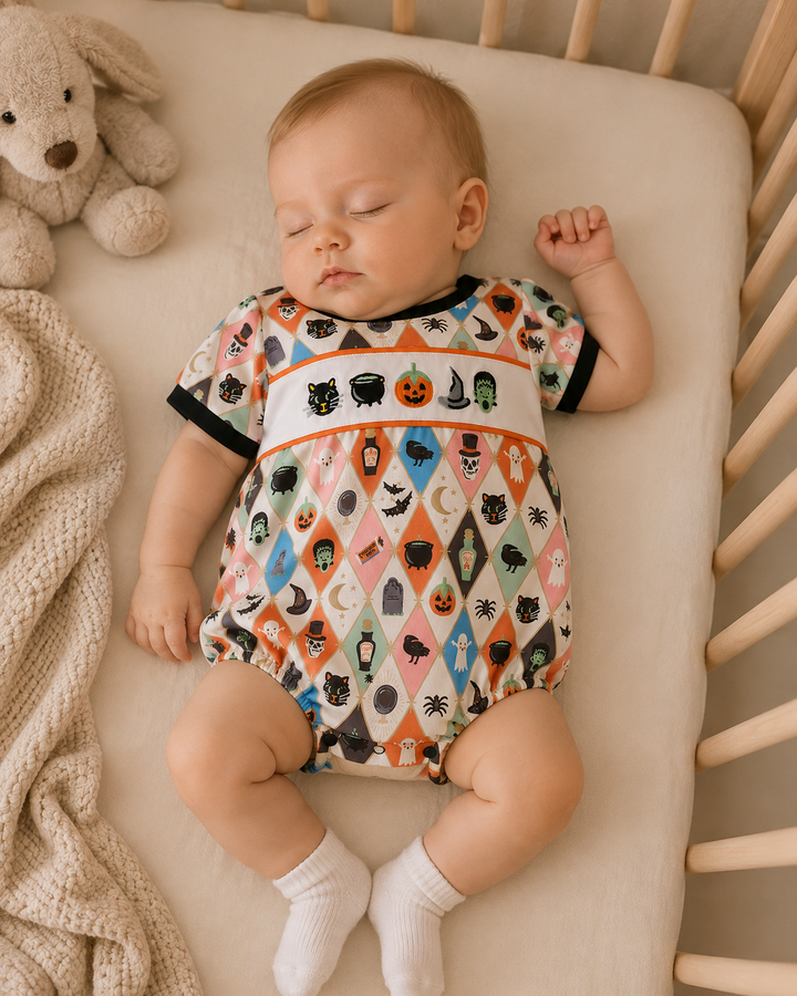 A sleeping baby lying on their side in a wooden crib, wearing a short-sleeve Halloween-themed bodysuit with a multicolor harlequin pattern of pumpkins, ghosts, bats, and moons, along with white socks.