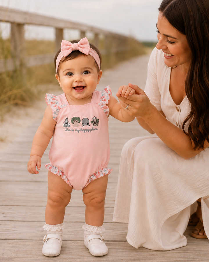 Baby in a pink outfit with ruffles and a bow, standing on a wooden boardwalk with a woman holding their hand.