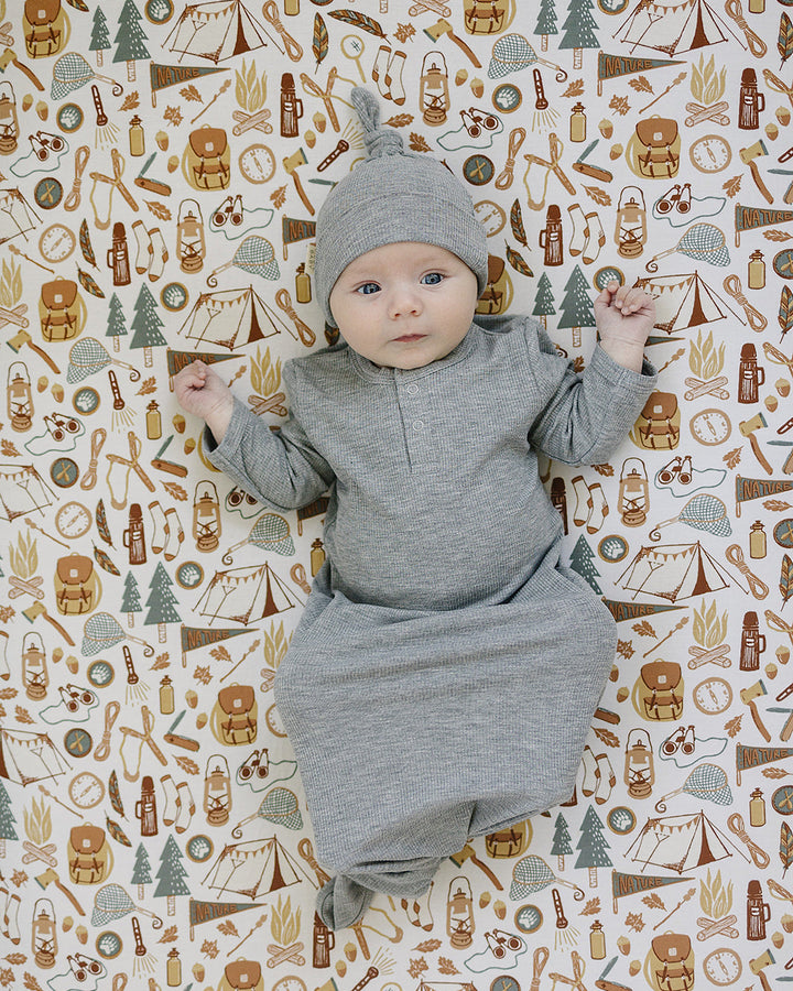 Baby in gray outfit lying on a patterned background with camping and nature-themed illustrations.