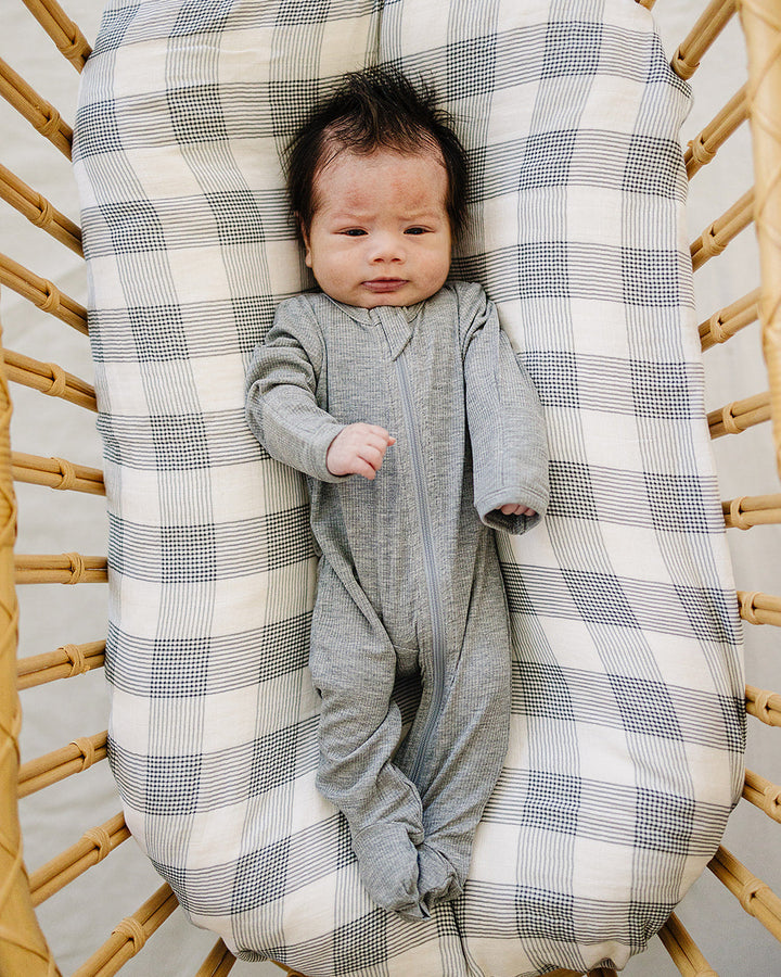 Baby in a gray onesie lying on a checkered blanket inside a wicker basket.
