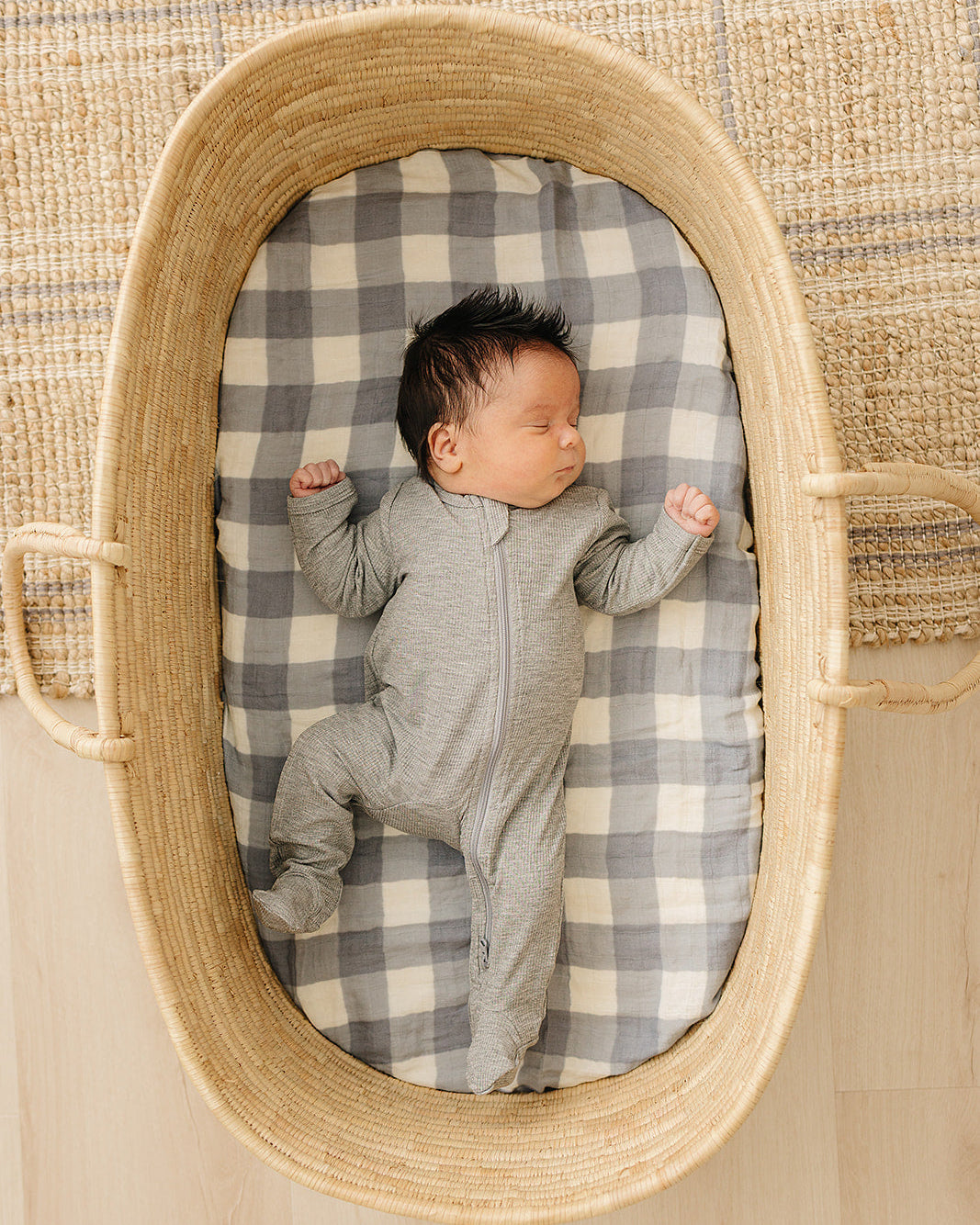 Newborn baby in a gray onesie lying on a checkered blanket inside a woven basket.