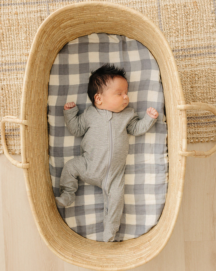 Newborn baby in a gray onesie lying on a checkered blanket inside a woven basket.