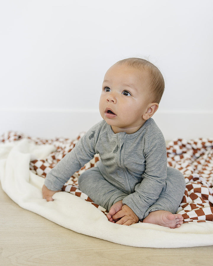 Baby in a gray outfit sitting on a white blanket with a patterned rug underneath.
