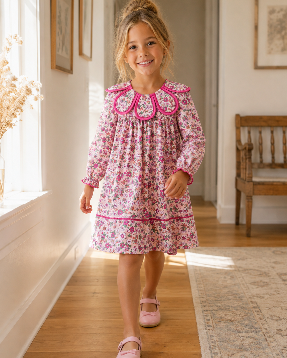 A young girl with blonde hair in a messy bun walks down a sunlit hallway wearing a pink floral long-sleeve dress with a ruffled Peter Pan collar and pink Mary Jane shoes.
