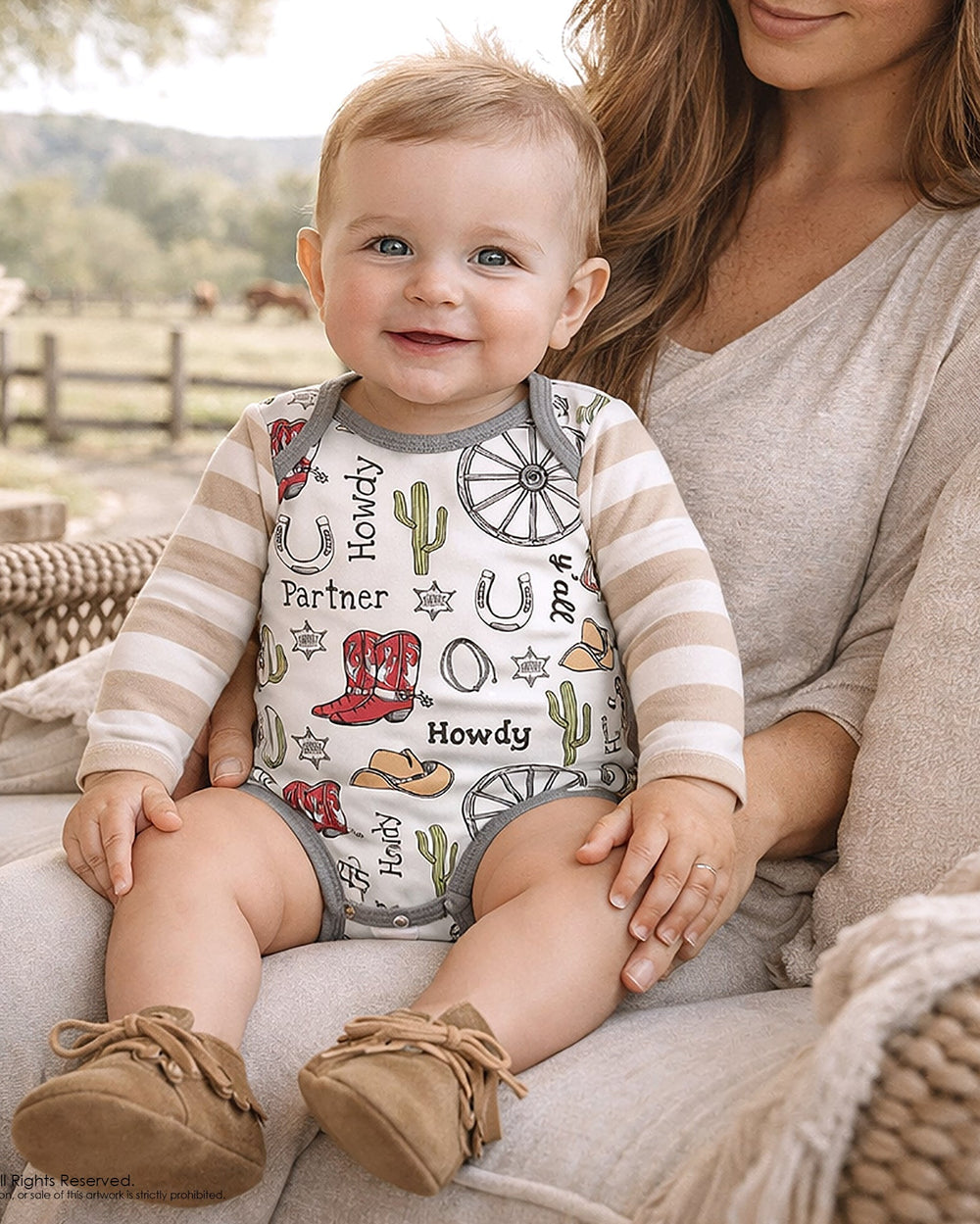 Baby wearing a 'Howdy' themed onesie sitting on a person's lap outdoors.