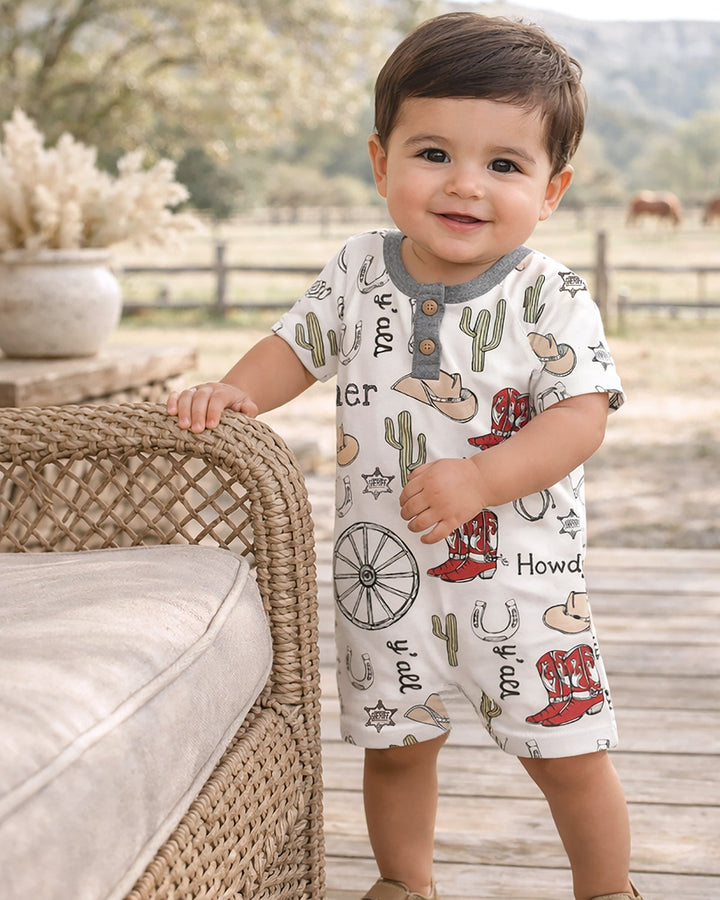 Child wearing a cowboy-themed romper standing on a wooden deck with a scenic background.