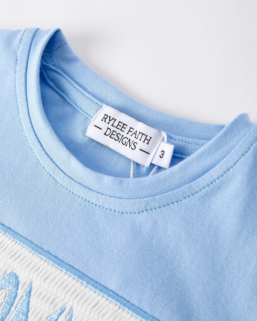 Close-up of a blue garment with a label from Rylee Faith Designs on a white background
