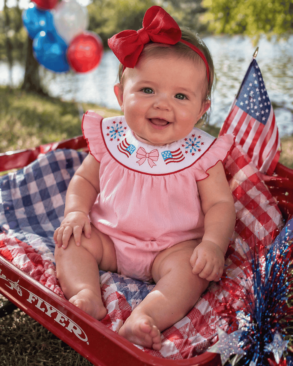 Baby in a red, white, and blue outfit with an American flag in a red wagon.