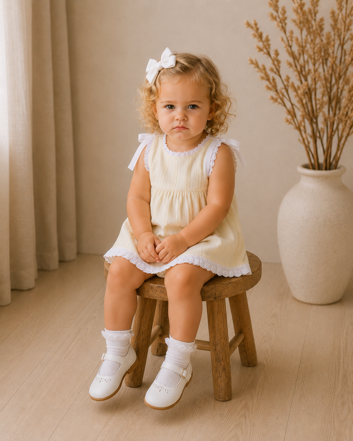 A toddler girl with curly blonde hair sits on a small wooden stool in a warm, neutral room. She is wearing a cream-colored sleeveless dress with white lace trim, white socks, and white Mary Jane shoes, and has a white bow in her hair.