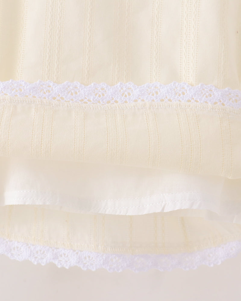Close-up of a cream-colored fabric garment with white lace trim and vertical embroidered details.