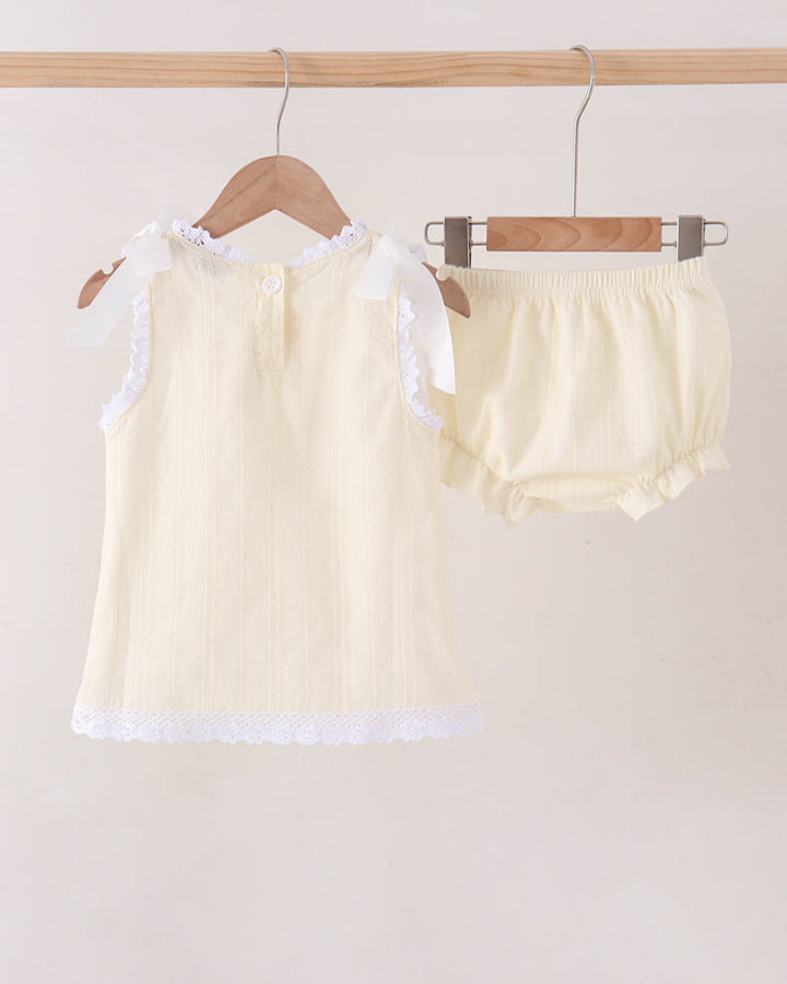 A two-piece baby outfit set consisting of a cream-colored sleeveless top with white lace trim and ruffle shoulder details, and matching cream bloomers, hanging on wooden hangers from a light wood clothing rack against a plain beige background.
