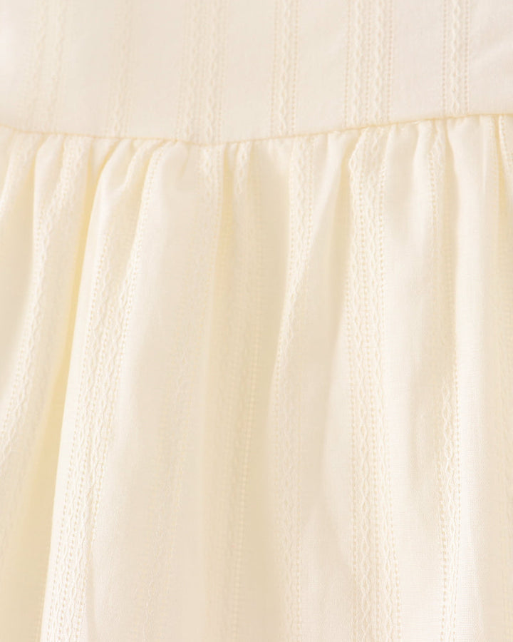 Close-up of a light cream or pale yellow dress fabric with vertical eyelet embroidery and a white lace trim at the neckline.