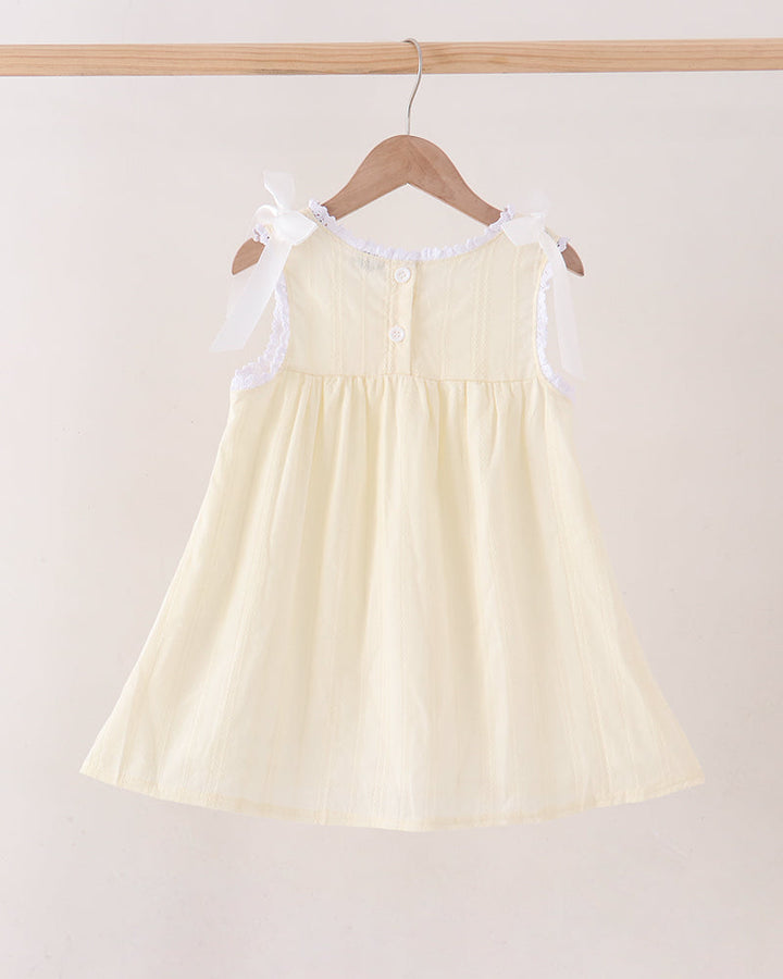 A pale yellow sleeveless baby dress with white lace shoulder straps and a ruffled neckline, hanging on a wooden hanger against a light beige background.