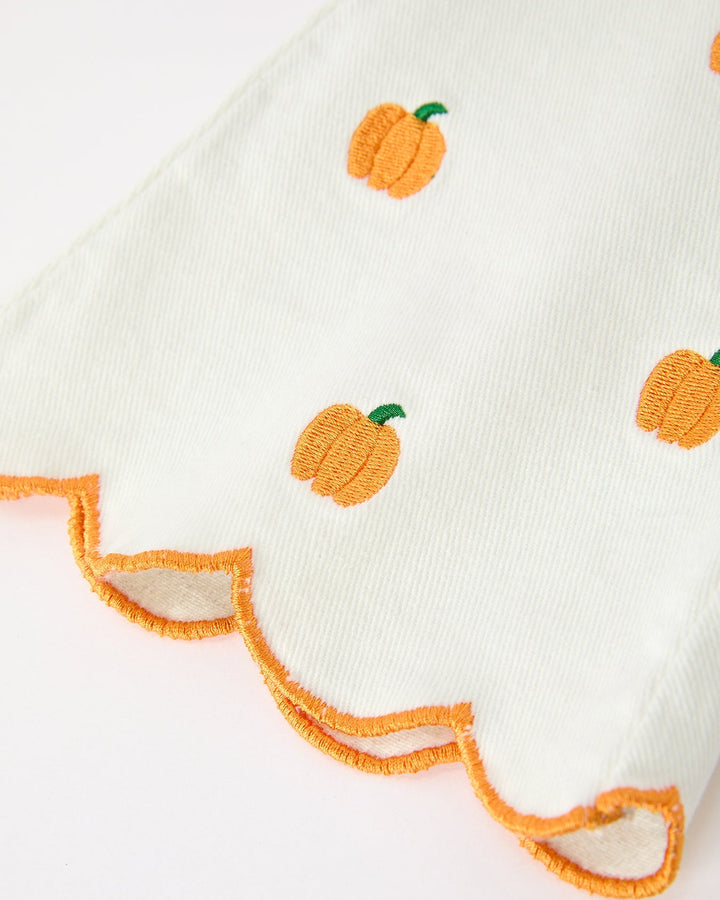 A close-up of a white fabric item, likely a baby blanket or towel, featuring embroidered orange pumpkins with green stems and a scalloped orange border.