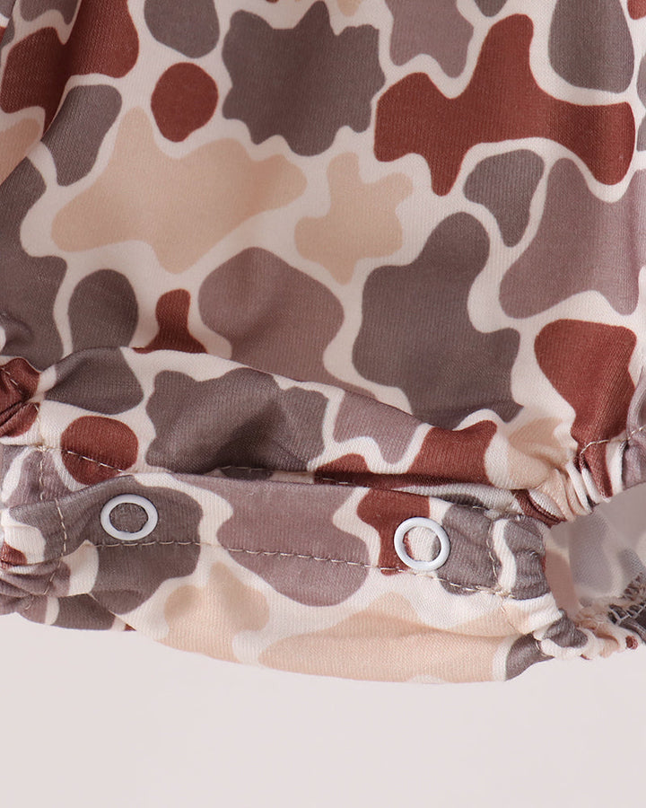 Close-up of a camouflage-patterned garment with brown, beige, and gray shapes outlined in white, featuring two silver metal snap buttons on a gathered elastic band.