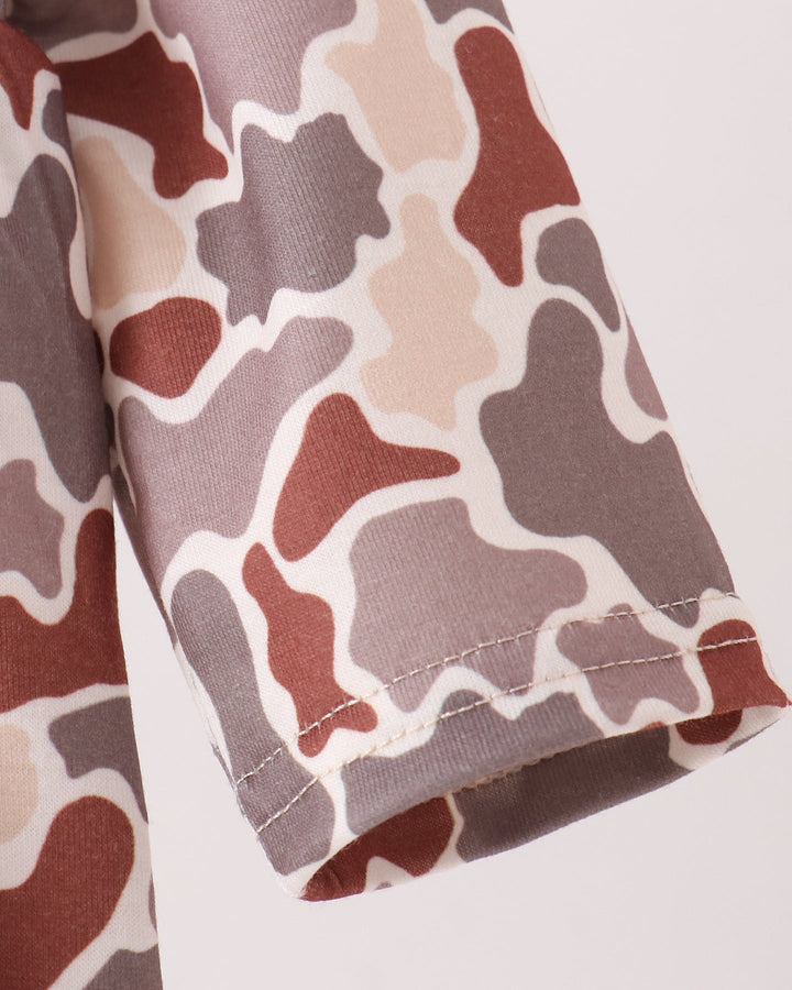 Close-up of a sleeve or fabric panel with an abstract camouflage-style pattern in muted brown, taupe, beige, and off-white, showing a folded hem and stitching on a light neutral background.
