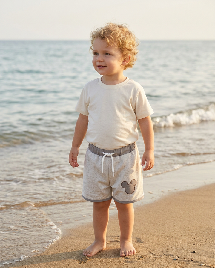 Child standing on a beach wearing a plain t-shirt and shorts with a mouse design.