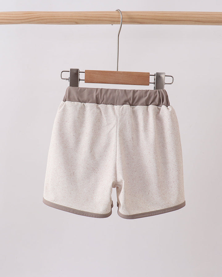 Beige swim shorts with brown trim hanging on a wooden hanger against a white background