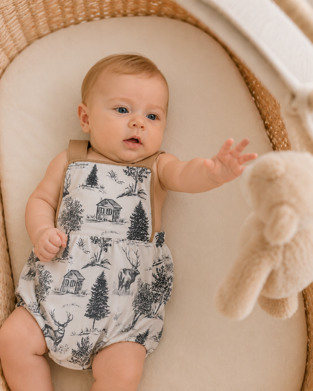 A baby wearing a white romper with a black toile de Jouy print of cabins, trees, and deer, lying in a wicker Moses basket next to a plush teddy bear.