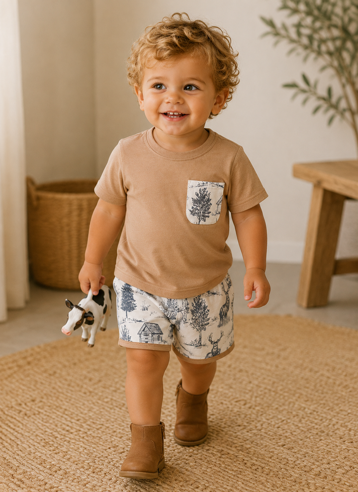 A toddler walking on a woven rug indoors, wearing a tan short-sleeve T-shirt with a blue tree-print chest pocket and matching blue-and-white patterned shorts, paired with tan ankle boots and holding a small cow toy.