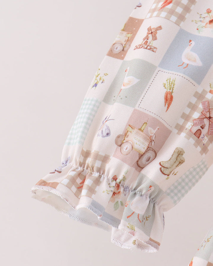 A close-up of a baby garment sleeve featuring a soft patchwork print with illustrations of carrots, a goose, a cart, and gingham checks in pastel tones, finished with a ruffled hem.
